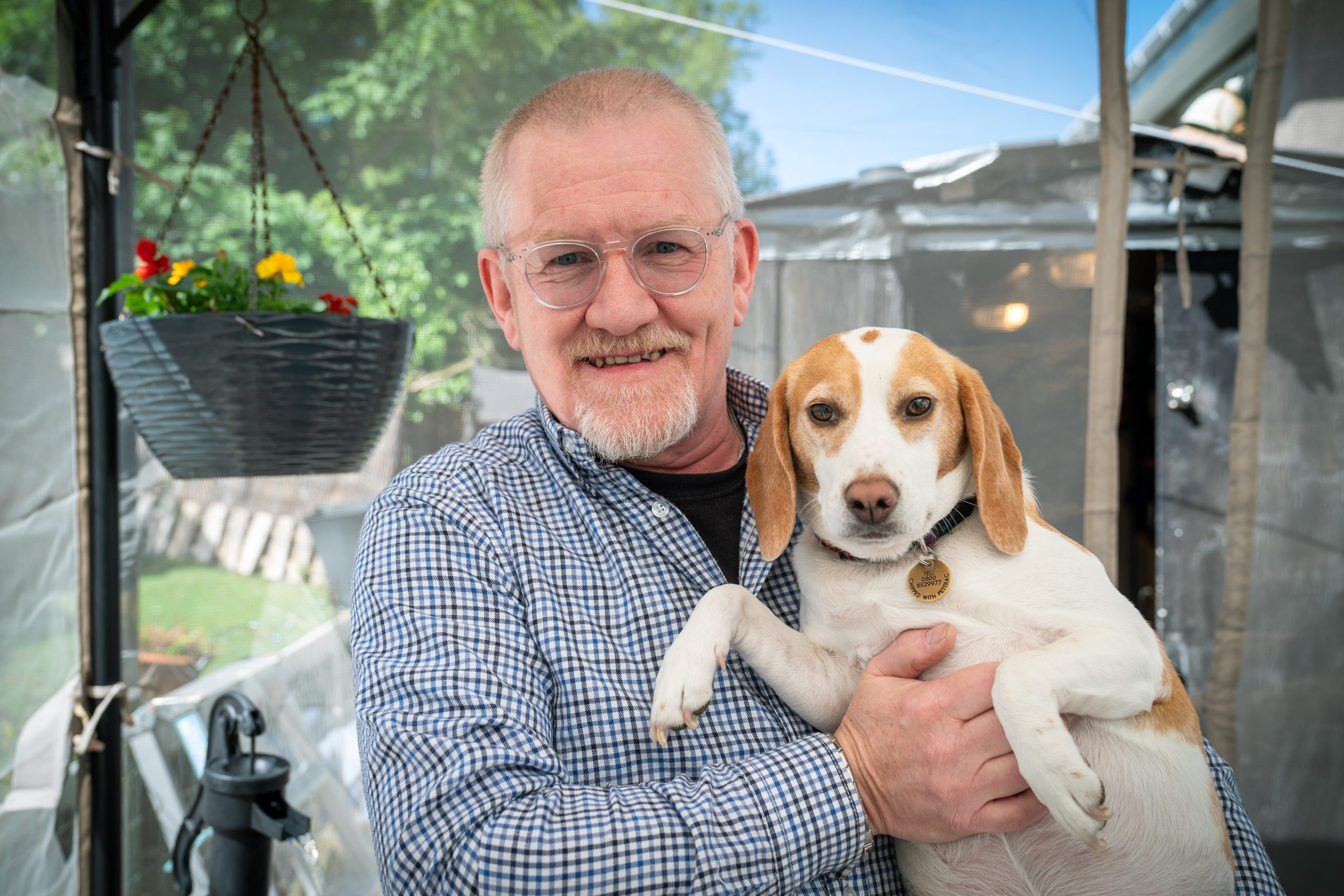 Trivallis Housing Landlord Wales An older man with glasses and a checked shirt smiles while holding a beagle. They are outside near a hanging flower basket, with greenery and a structure visible in the background.