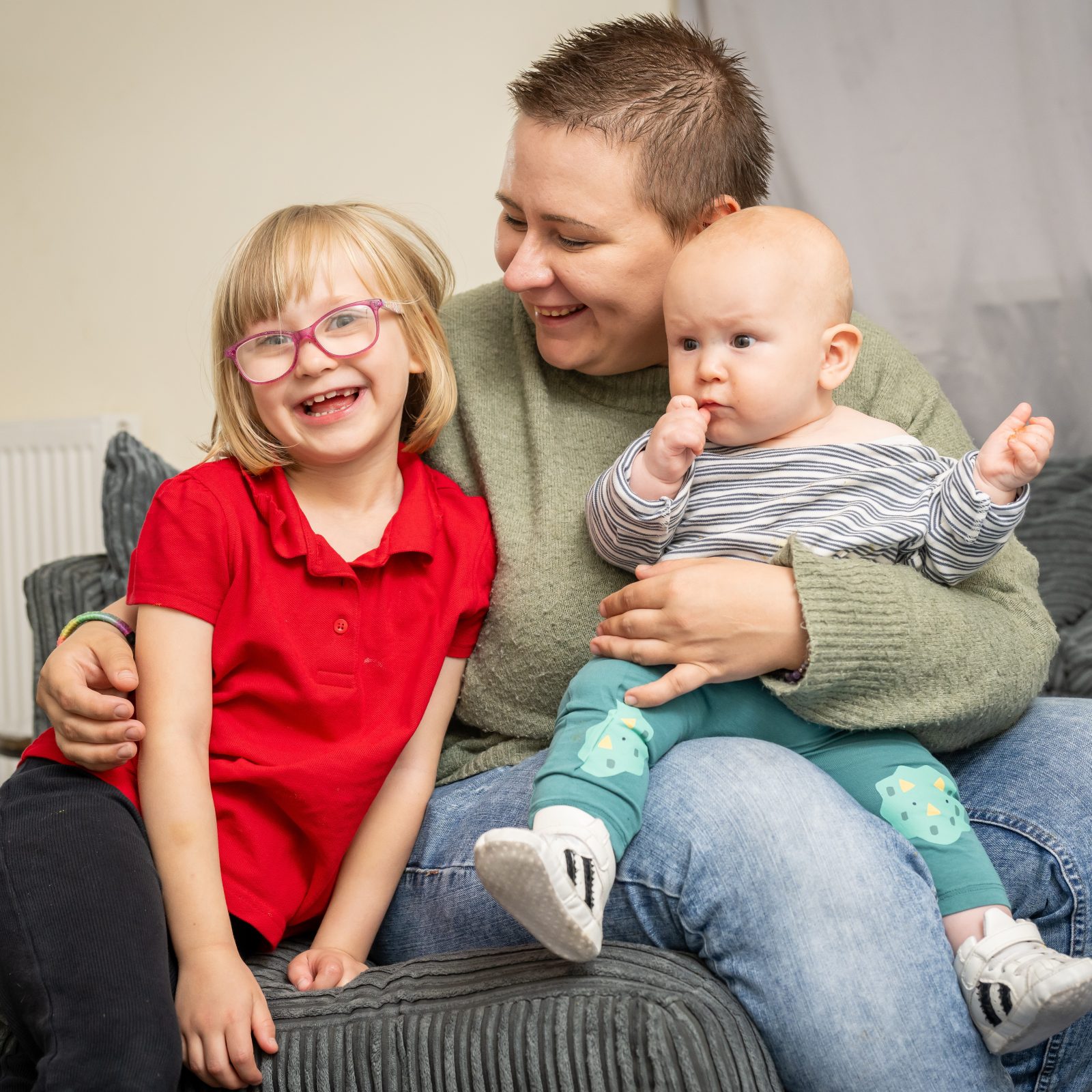 Trivallis Housing Landlord Wales A woman with short hair sits on a couch holding a baby, while a young girl with glasses and a red shirt sits beside them, all indoors with a grey curtain in the background.