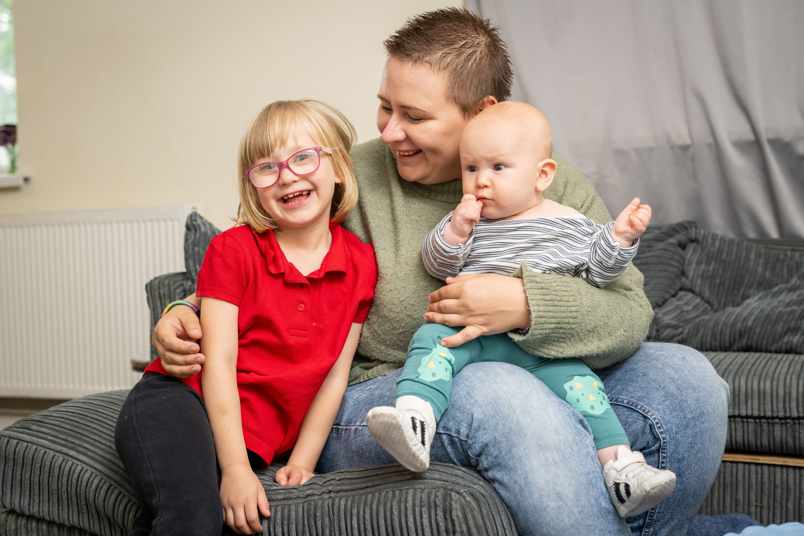 Trivallis Housing Landlord Wales A woman with short hair sits on a couch holding a baby, while a young girl with glasses and a red shirt sits beside them, all indoors with a grey curtain in the background.