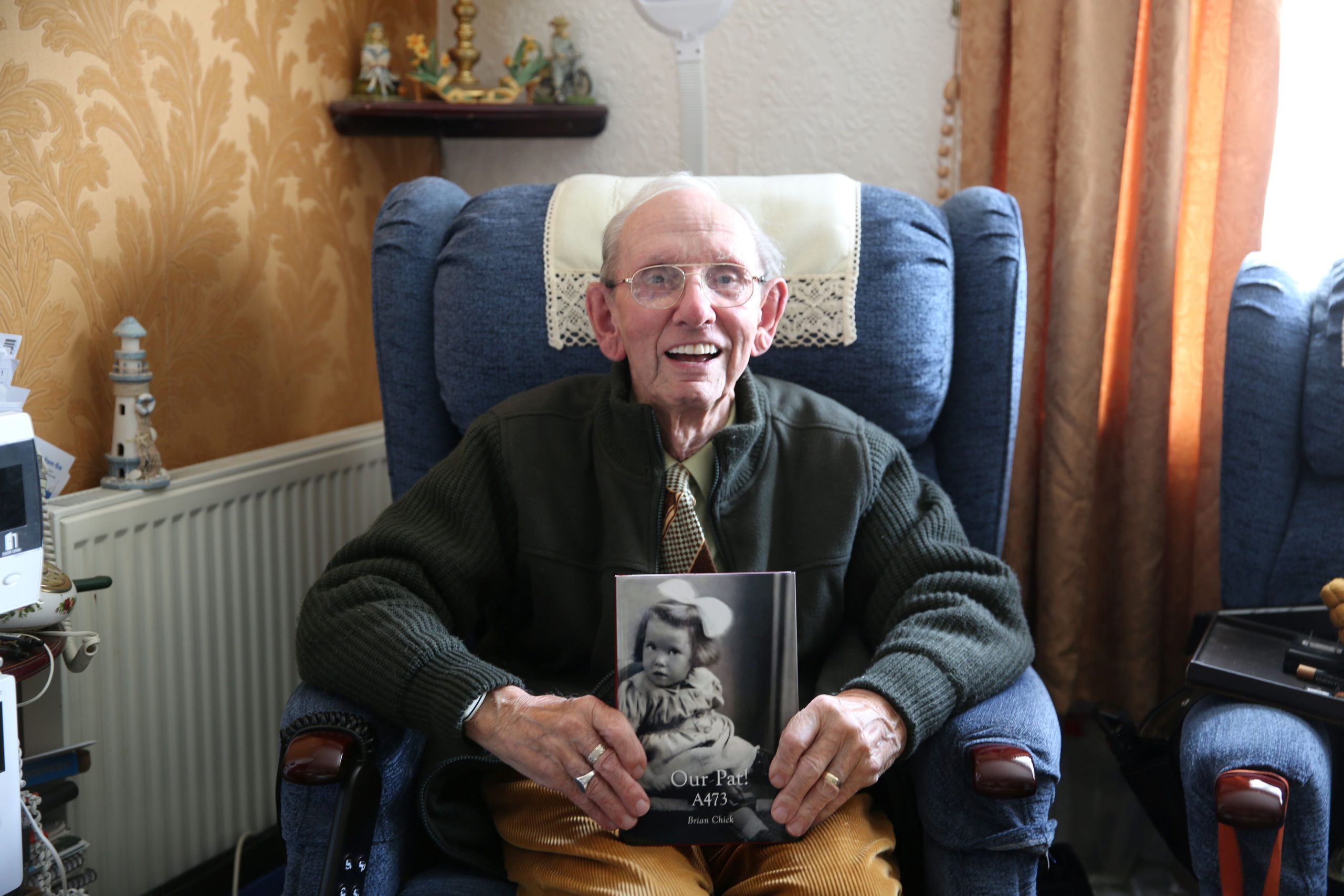 Trivallis Housing Landlord Wales An elderly man with glasses sits in a blue armchair, smiling and holding a book with a black-and-white photo of a child on the cover. The room has ornaments on a shelf and golden curtains by the window.