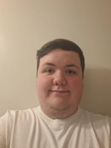 Trivallis Housing Landlord Wales A person with short dark hair and fair skin is facing the camera and smiling slightly. They are wearing a plain white t-shirt and are standing in front of a beige wall.