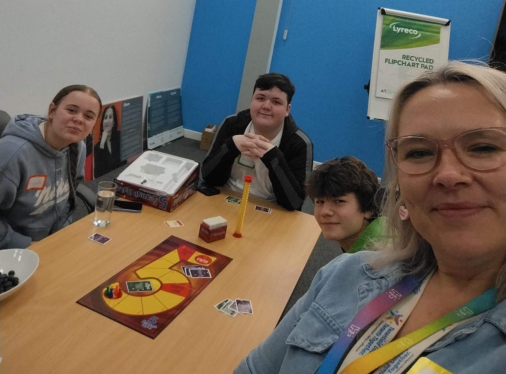 Trivallis Housing Landlord Wales Four people sit around a table playing a board game, with cards and pieces visible. One person in the foreground is taking a selfie. The room has blue walls, large windows, and office supplies in the background.