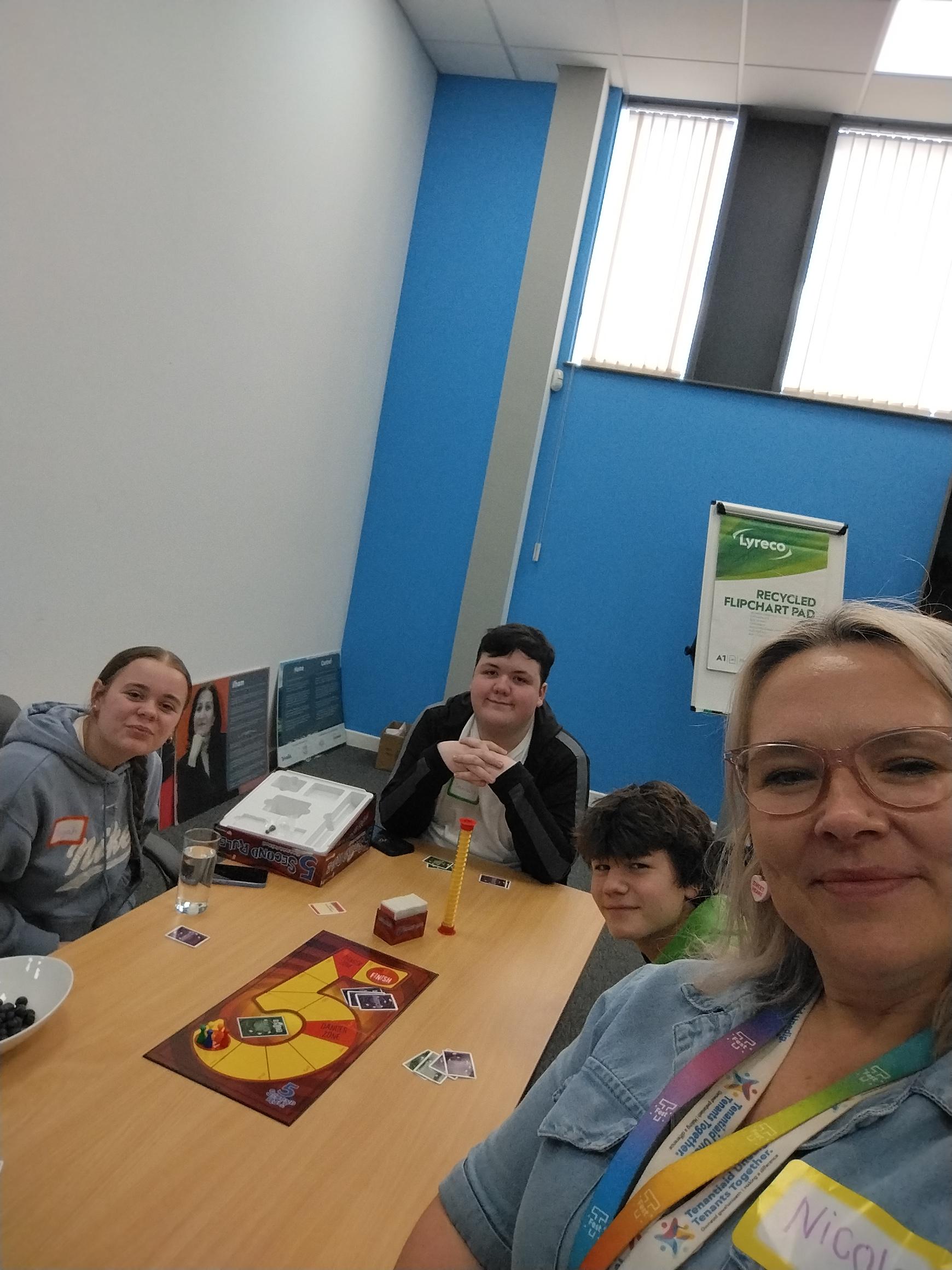 Trivallis Housing Landlord Wales Four people sit around a table playing a board game, with cards and pieces visible. One person in the foreground is taking a selfie. The room has blue walls, large windows, and office supplies in the background.