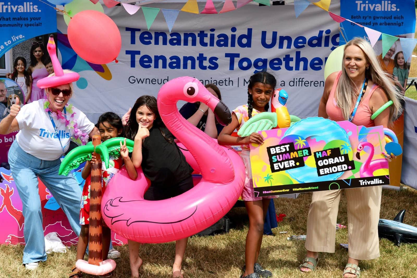 Trivallis Housing Landlord Wales A group of children and two adults pose outdoors with inflatable pool toys and a colorful sign at a Trivallis Tenants Together event, with a banner and decorations in the background.
