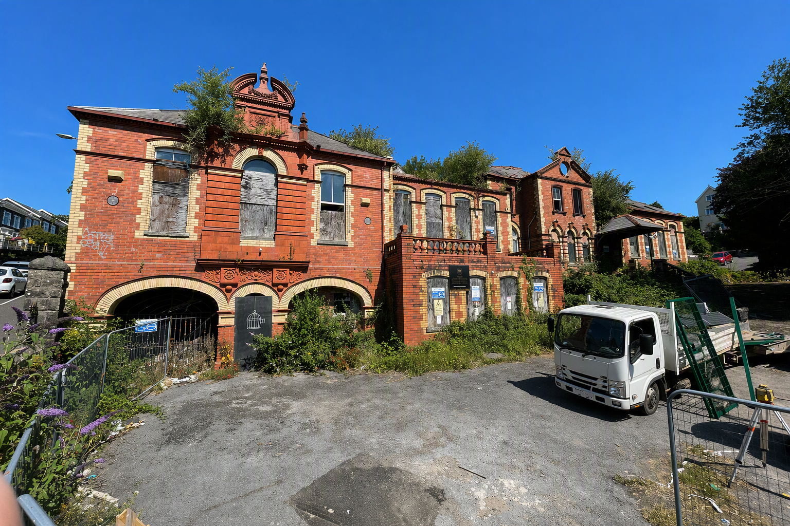 Trivallis Housing Landlord Wales Merthyr General Hospital, a rundown red-brick building with boarded-up windows and overgrown vegetation sits beside a vacant lot. A white truck is parked nearby under a clear blue sky.