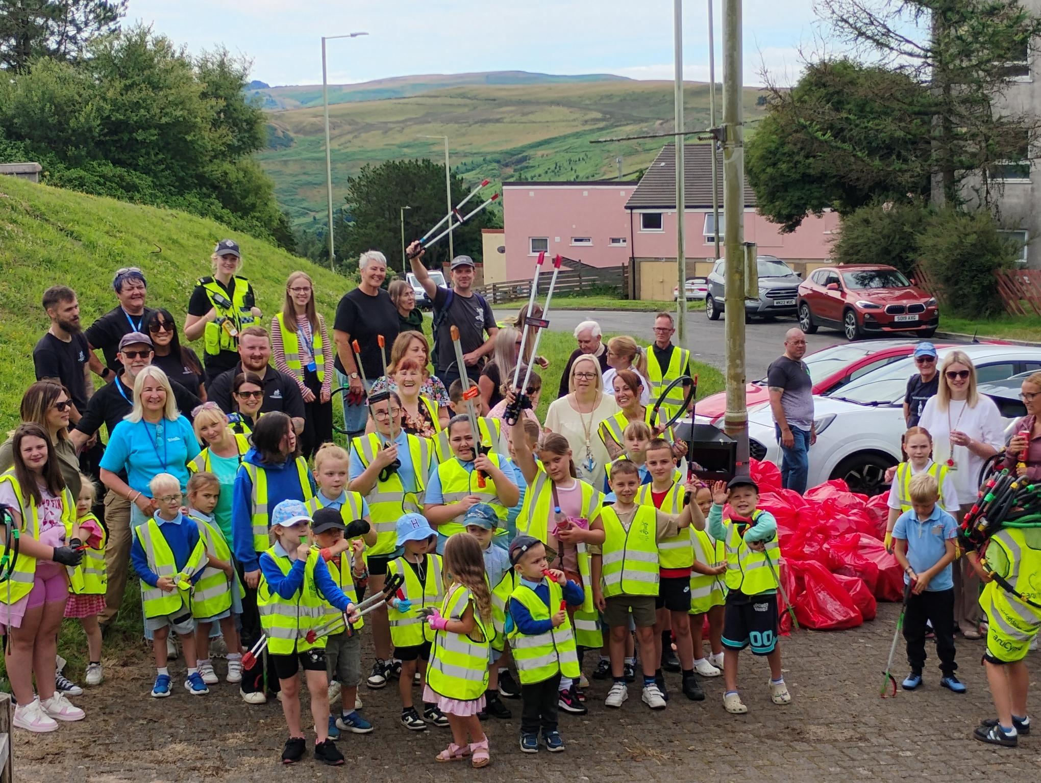 Trivallis Housing Landlord Wales A large group of adults and children in yellow safety vests poses outdoors with litter pickers and filled trash bags, in front of parked cars and grassy hills under a cloudy sky.