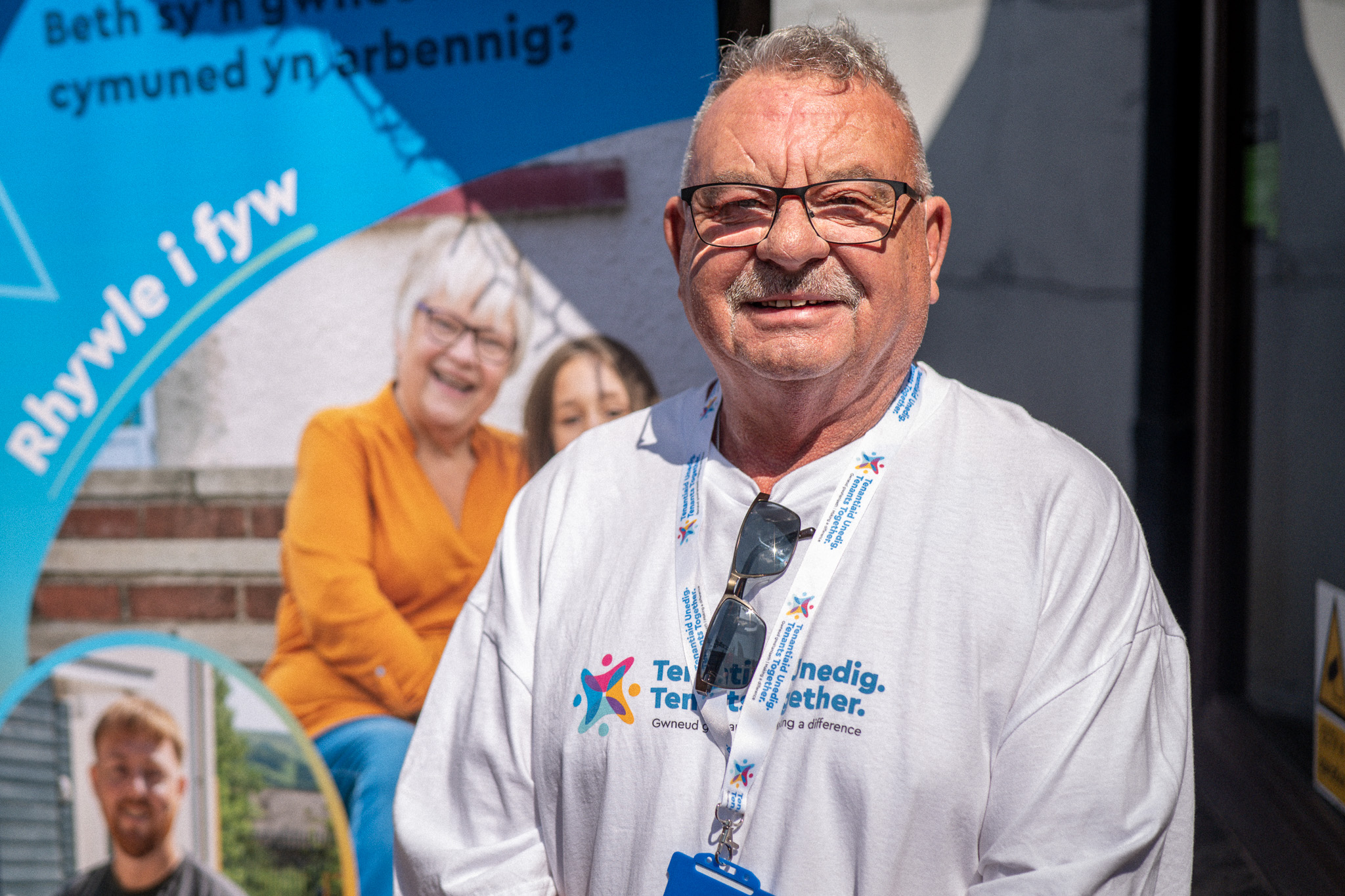 Trivallis Housing Landlord Wales An older man with glasses and a white “Ten Together” T-shirt stands outdoors, smiling. He wears a lanyard and is in front of a blue banner with Welsh text and photos of people in the background.