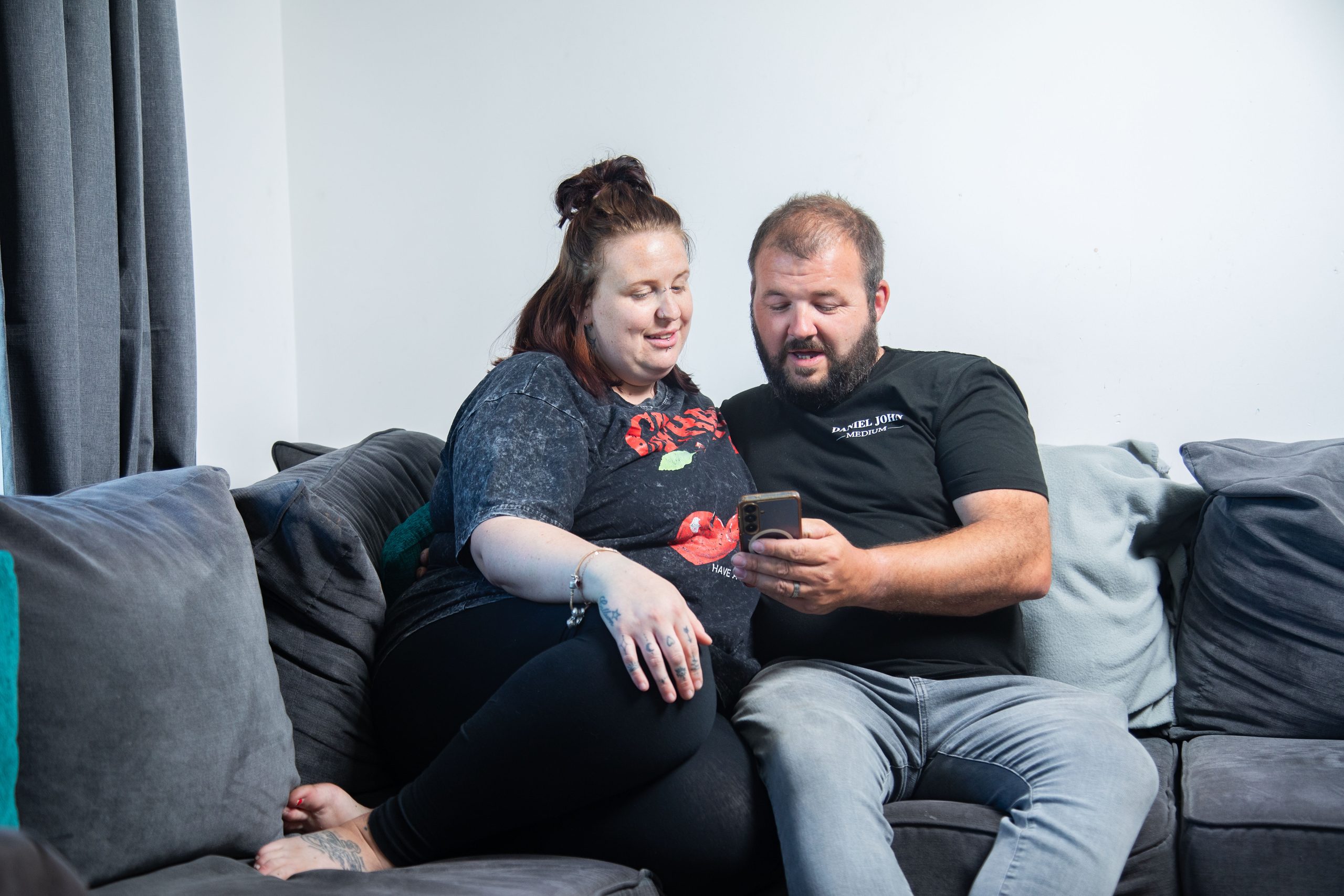 Trivallis Housing Landlord Wales A woman and a man sit closely together on a grey sofa, looking at a mobile phone the man is holding. The background includes light-coloured walls and curtains. Both appear relaxed and engaged with the phone.