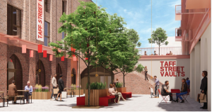 Trivallis Housing Landlord Wales People sit, walk, and talk in a modern outdoor courtyard with brick walls, trees, benches, and red signage that reads “TAFF STREET VAULTS.” Some people are on an elevated terrace, and red accents decorate the area.