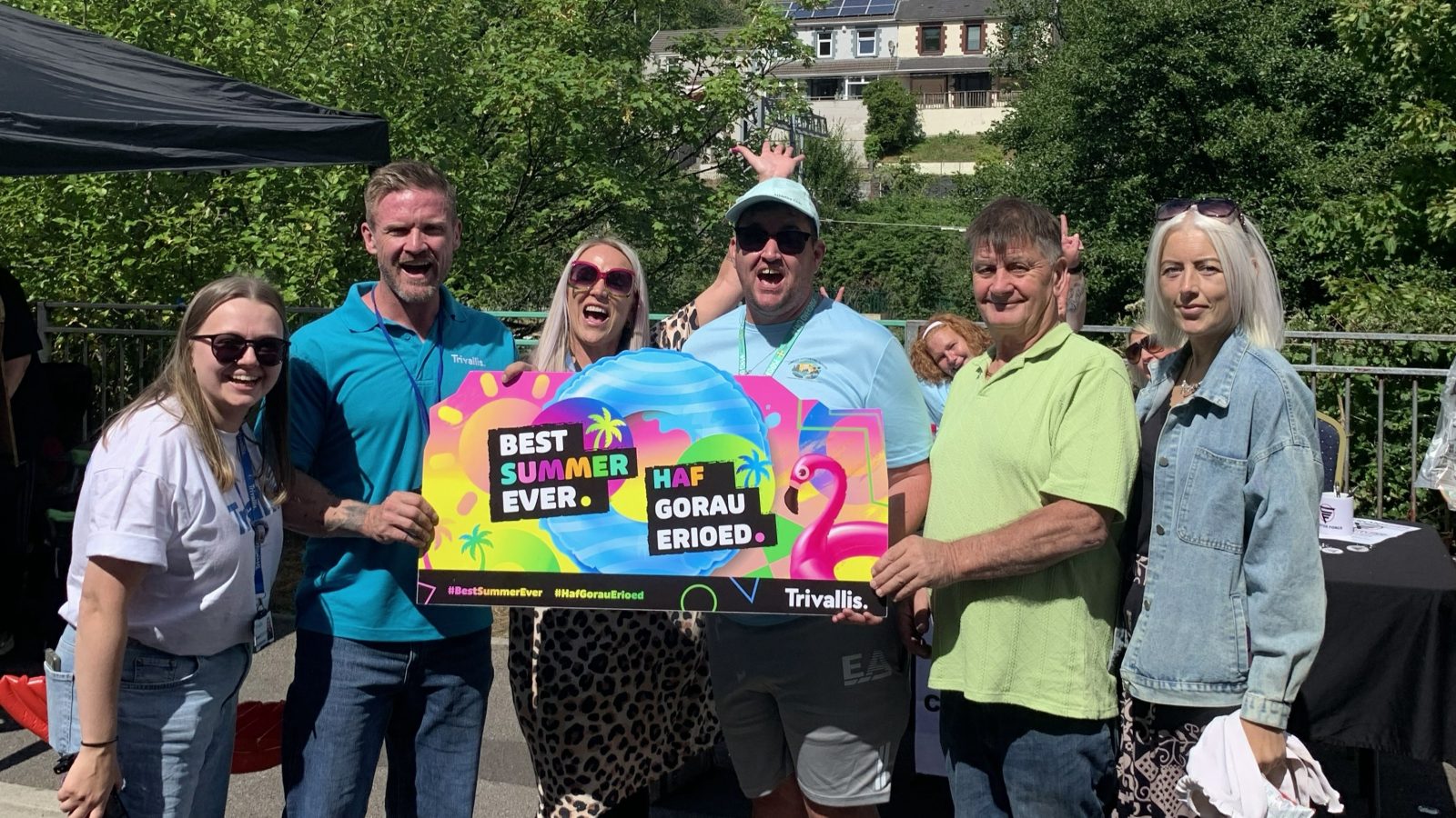 Trivallis Housing Landlord Wales A group of six people smiling outdoors while holding a sign that says “Best Summer Ever” and “Haf Gorau Erioed” with a flamingo and beach-themed design.