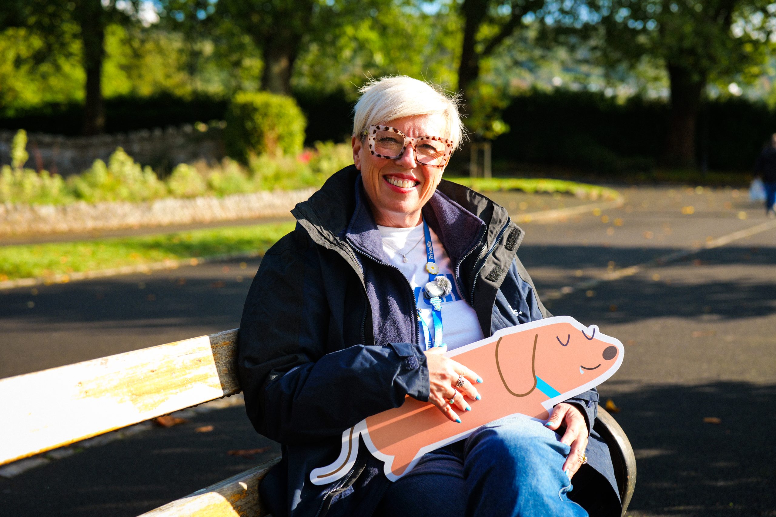 Trivallis Housing Landlord Wales A smiling woman with short blonde hair and glasses sits on a park bench holding a flat, illustrated cut-out of a dachshund.